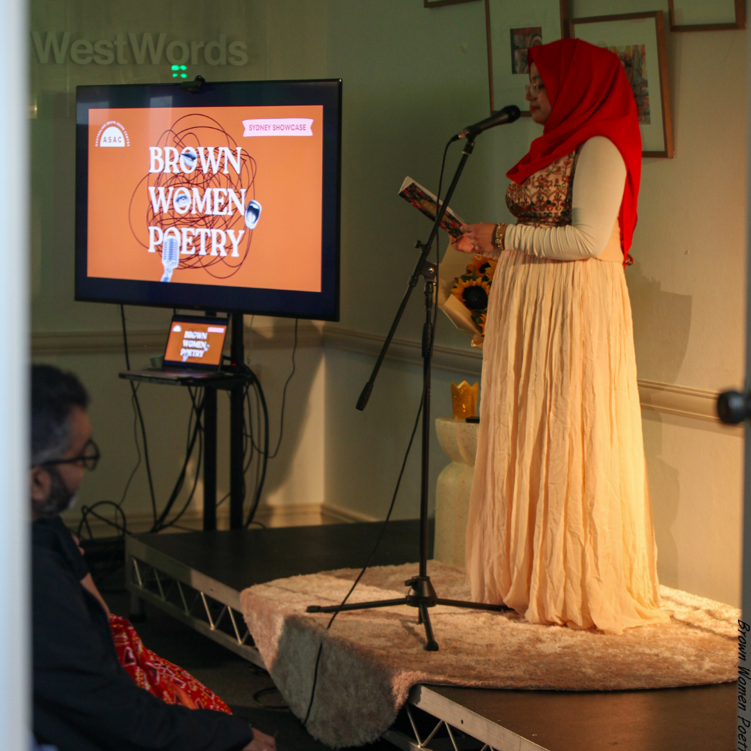 A woman in a red hijab reads poetry on stage beside a screen displaying ‘Brown Women Poetry’ at a Sydney showcase.